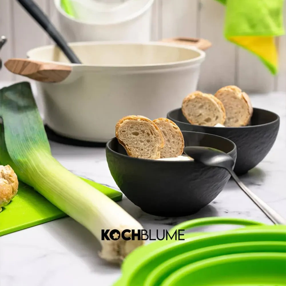 Zwei schwarze Suppenschüsseln mit Brotscheiben, ein Lauch, ein Topf und grüne Küchenutensilien auf einer Arbeitsplatte.