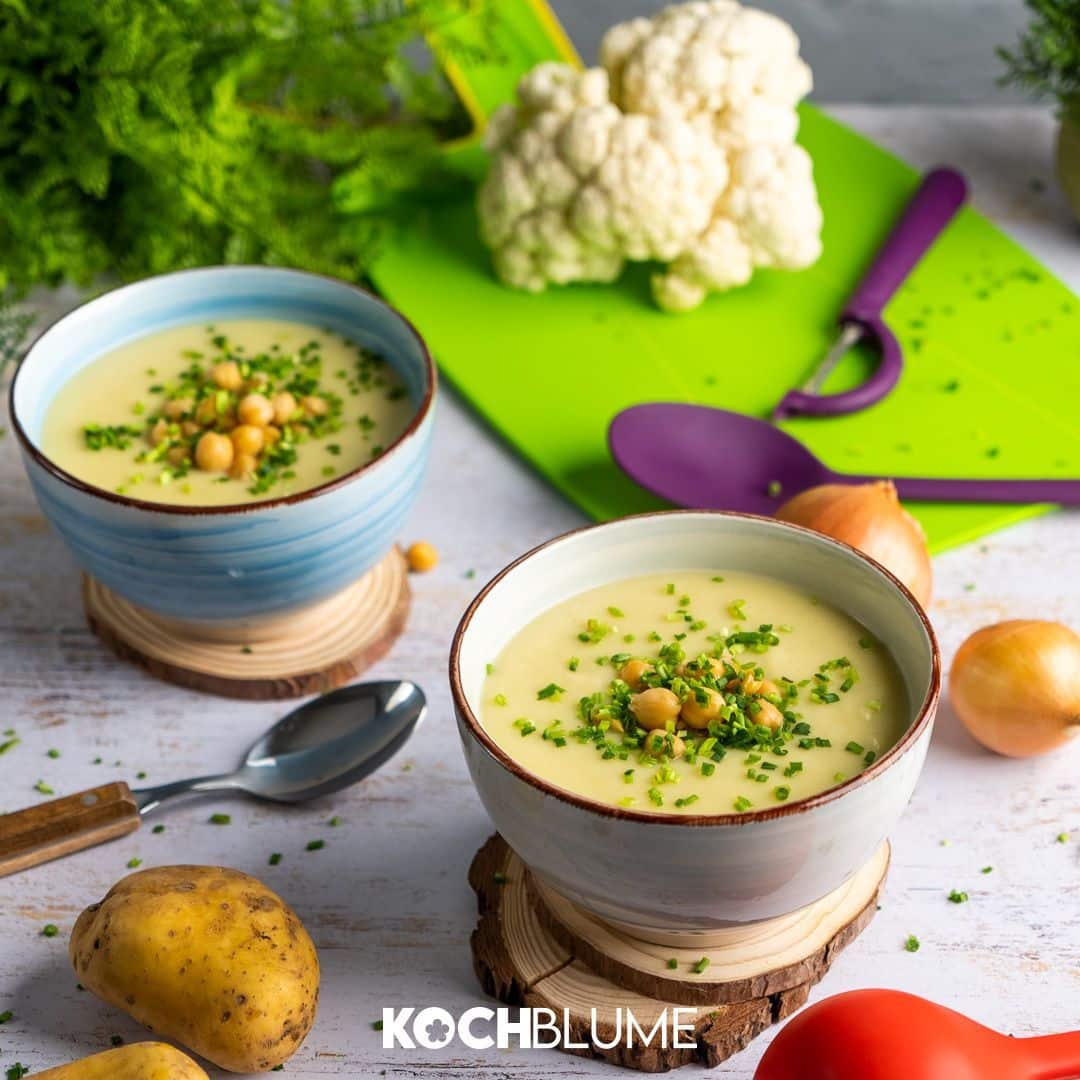 Zwei Schüsseln mit cremiger Suppe, garniert mit Kichererbsen und Schnittlauch, umgeben von frischem Gemüse und Besteck.