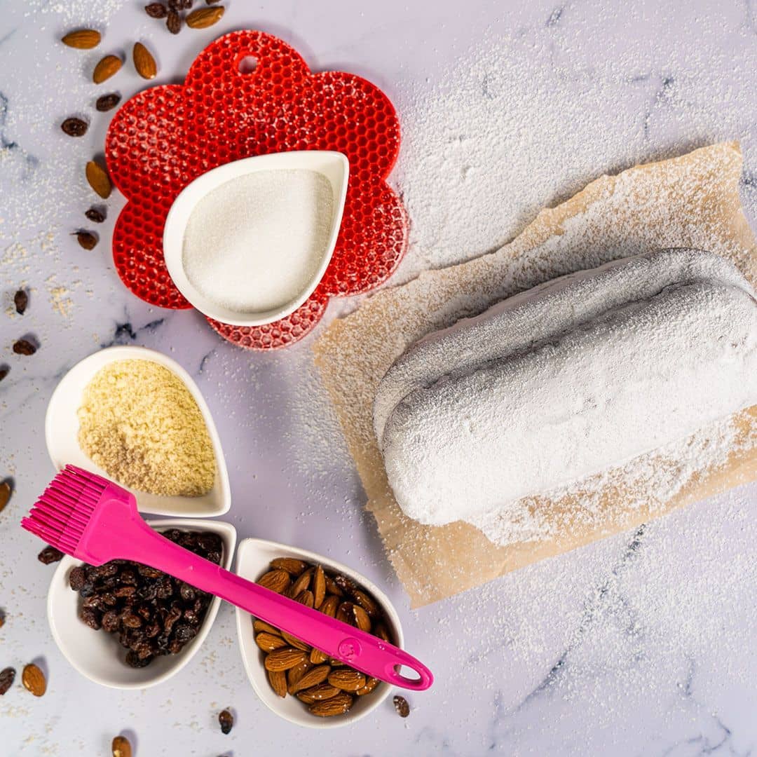 Ein mit Puderzucker bestäubtes Brot, Nüsse, Rosinen und Zucker in Schalen auf einer Marmorplatte mit einem rosa Pinsel.
