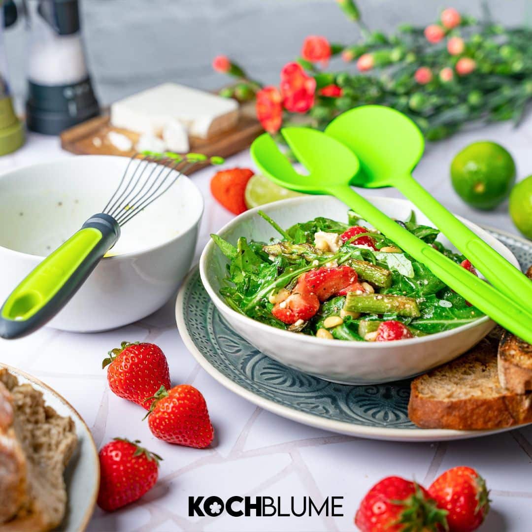 Frischer Salat mit Tomaten und Spargel in einer Schüssel, grünes Besteck, Brot, Erdbeeren und Limette auf dem Tisch.