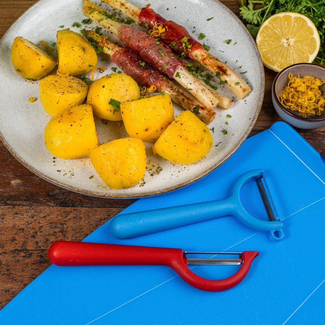 Teller mit gekochten Kartoffeln, in Fleisch gewickeltem weißem Spargel, Zitronenhälften, Zitronenschale, Kräutern und Schälmessern auf einer blauen Unterlage.