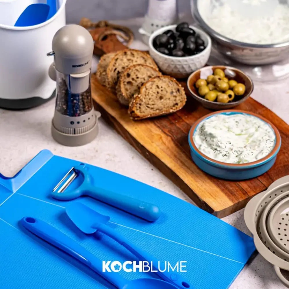 Auf einem Tisch mit einem blauen Schneidebrett liegen geschnittenes Brot, Oliven, Dip und Küchenutensilien.