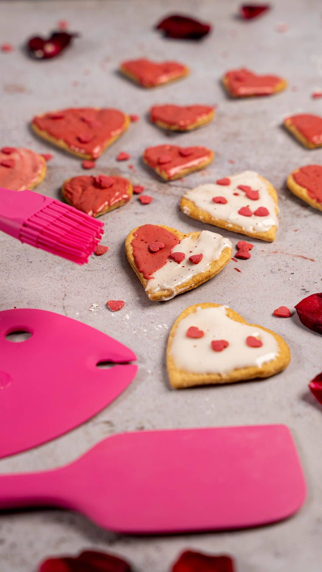 Herzförmige Plätzchen mit rot-weißer Zuckerglasur, einem rosa Spatel und einem Pinsel auf grauem Untergrund.