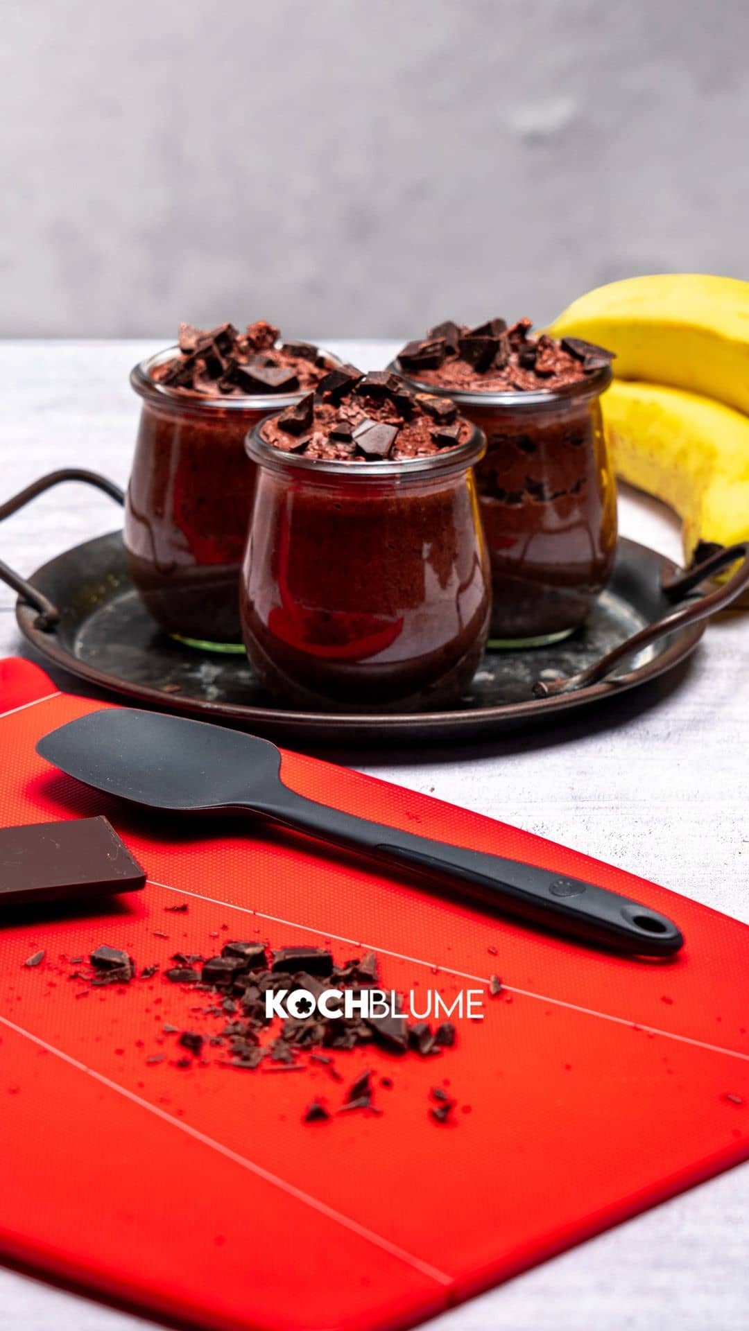 Drei Gläser Schokoladenmousse mit Schokoladenstückchen, Bananen, einem Spatel und einer Tafel Schokolade auf einem roten Brett.