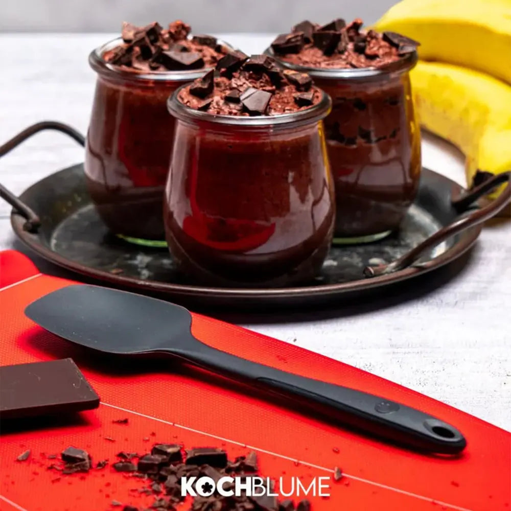 Drei Gläser mit Schokoladenmousse und Schokoladenstückchen, ein Spatel, eine Banane und Schokolade auf einem roten Brett.