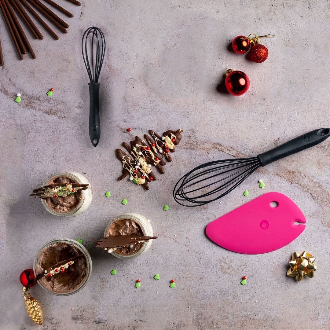 Zwei Schneebesen, ein rosa Pfannenwender, Weihnachtsleckereien und Dessertgläser auf einer hellen Unterlage mit weihnachtlicher Dekoration.