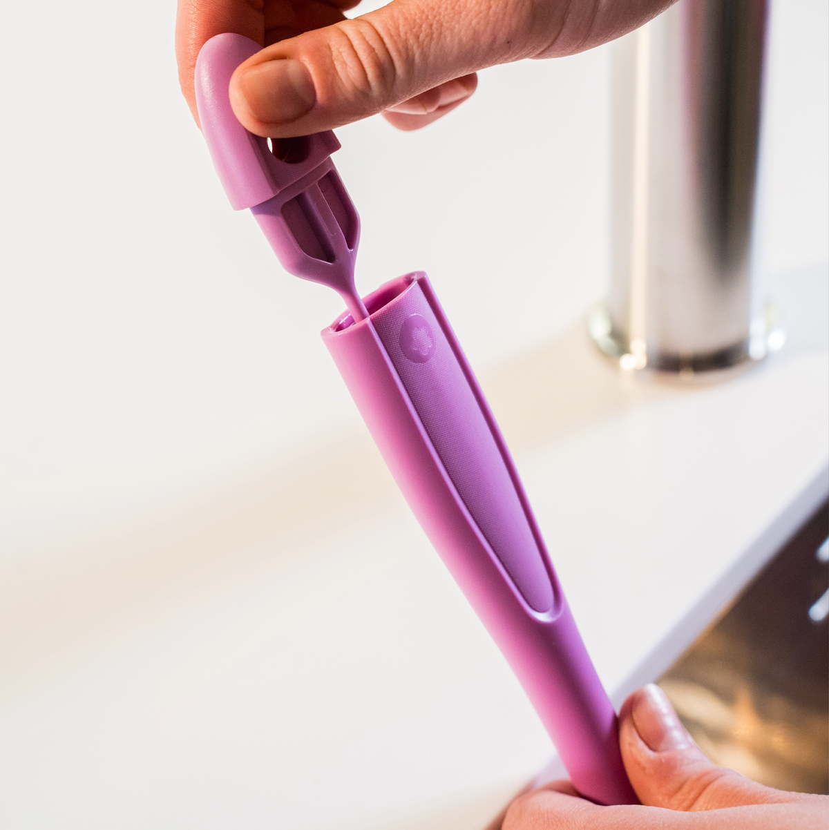 Eine Hand öffnet eine violette Silikonhülle der Marke „Becherbürste“ von Kochblume GmbH und gibt den Blick auf ein kleineres, dazu passendes Werkzeug frei – ideal für die Flaschenreinigung oder als praktische Trinkhalmbürste –, das in der Nähe eines Waschbeckens aufbewahrt wird.
