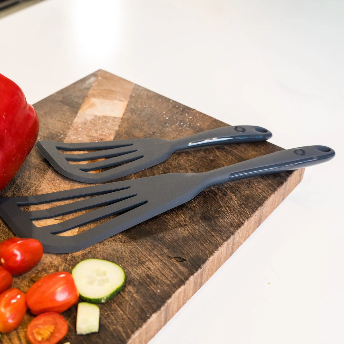 Zwei schwarze Design-Eckwender-Pfannenwender der Kochblume GmbH liegen auf einem Holzschneidebrett, umgeben von frischen, farbenfrohen Zutaten – darunter Tomatenscheiben, Gurke und eine rote Paprika –, was diese Küchenszene ideal für alle macht, die gesunde Mahlzeiten mit hochwertigen Küchenutensilien zubereiten möchten.