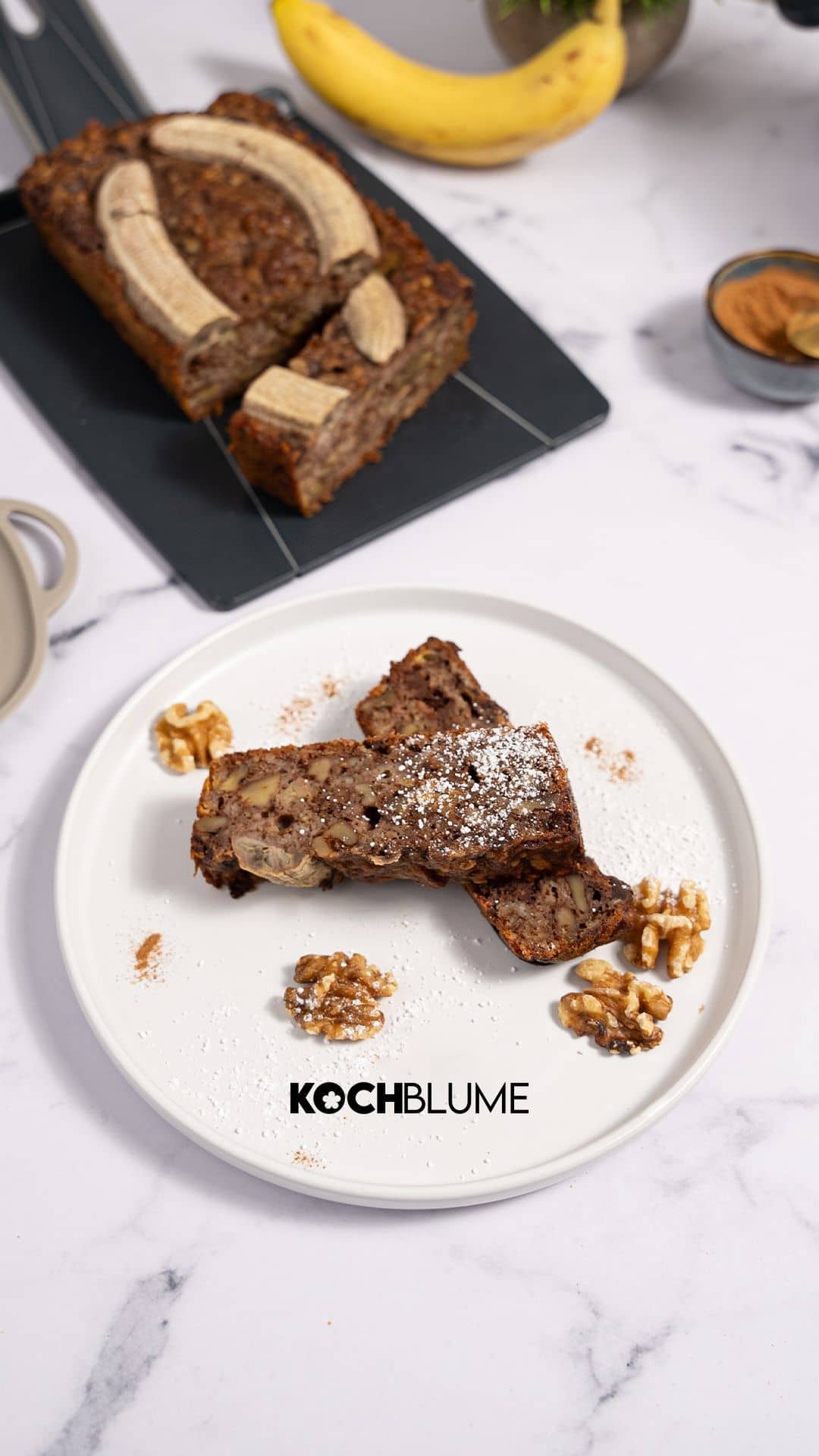 Zwei Scheiben Bananenbrot mit Walnüssen auf einem weißen Teller; im Hintergrund ein Laib Brot und eine Banane auf einer Marmorplatte.