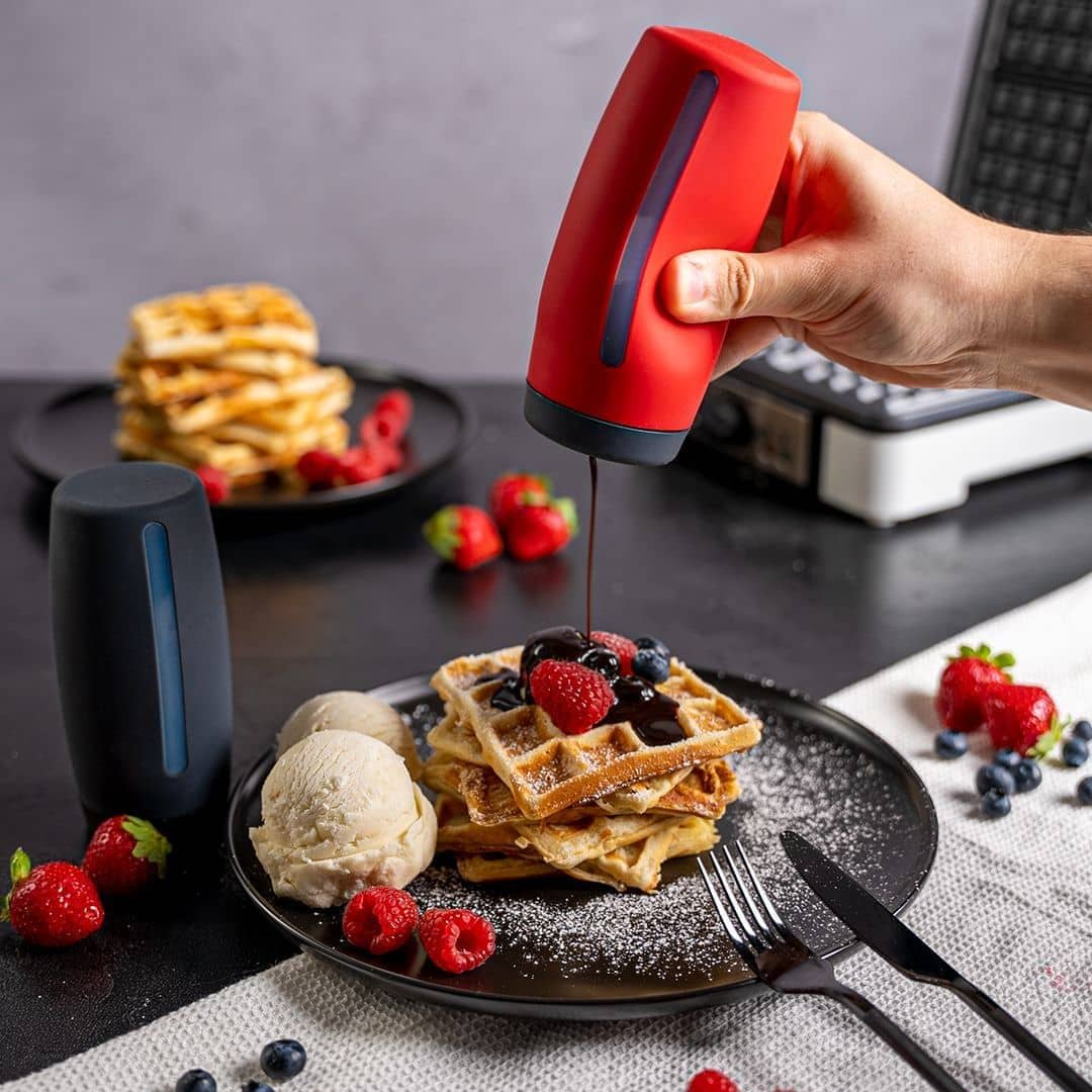 Ein Dosierspender der Firma Kochblume GmbH gibt Schokoladensoße auf Waffeln mit Beeren und Eis, im Hintergrund sind frische Beeren und Waffeln zu sehen.