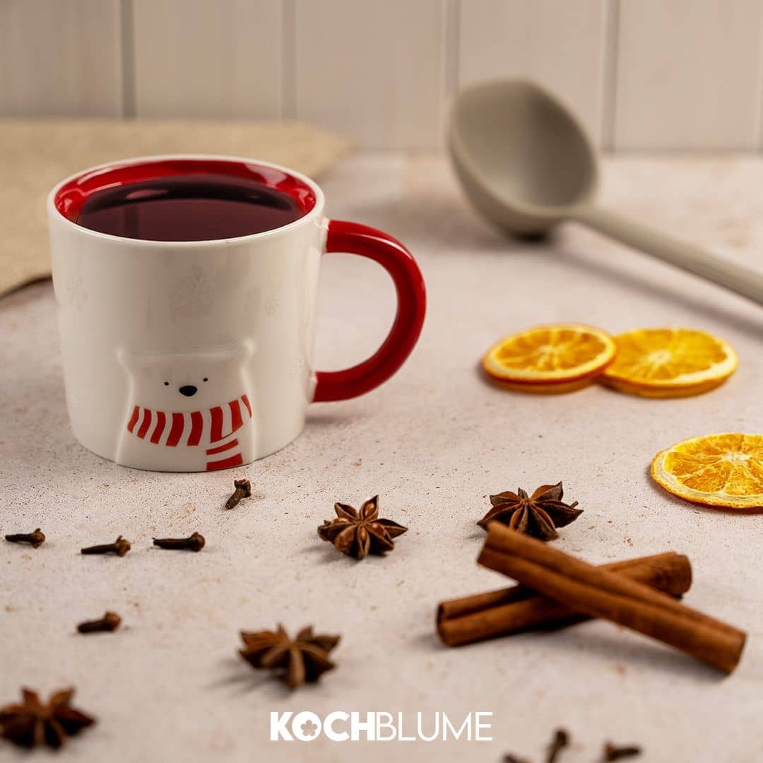 Eine weiße Tasse mit rotem Rand, Sternanis, Zimtstangen, getrockneten Orangenscheiben und einem grauen Löffel auf einem Tisch.