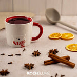 Eine weiße Tasse mit rotem Rand, Sternanis, Zimtstangen, getrockneten Orangenscheiben und einem grauen Löffel auf einem Tisch.