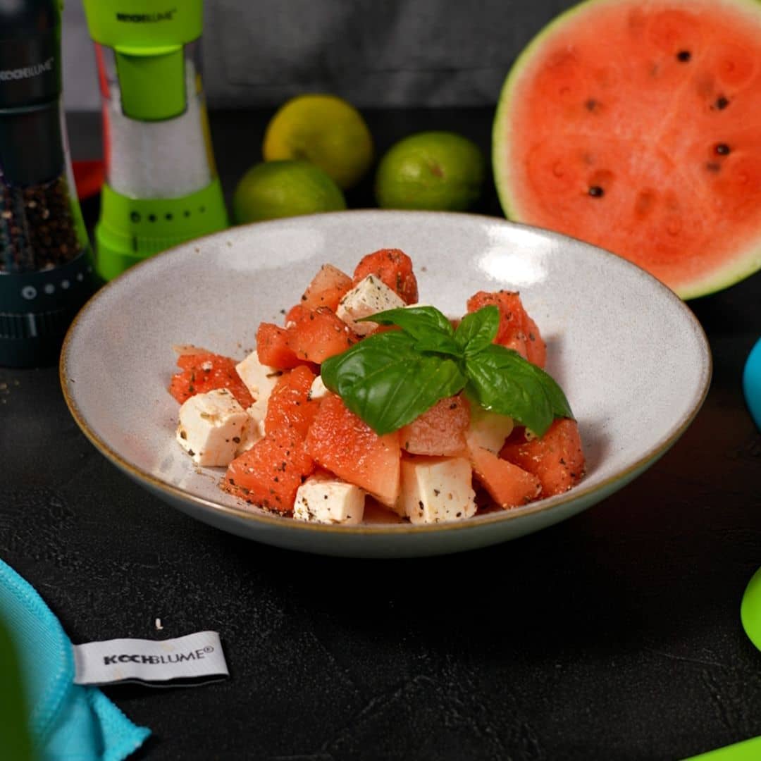 Eine Schüssel mit Wassermelonen-Feta-Salat, garniert mit Basilikum, im Hintergrund sind Wassermelonen und Limetten zu sehen.