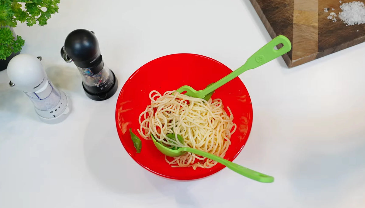 Eine rote Schüssel mit einfachen Spaghetti und grünem Besteck, daneben Salz- und Pfefferstreuer auf einer weißen Arbeitsplatte.