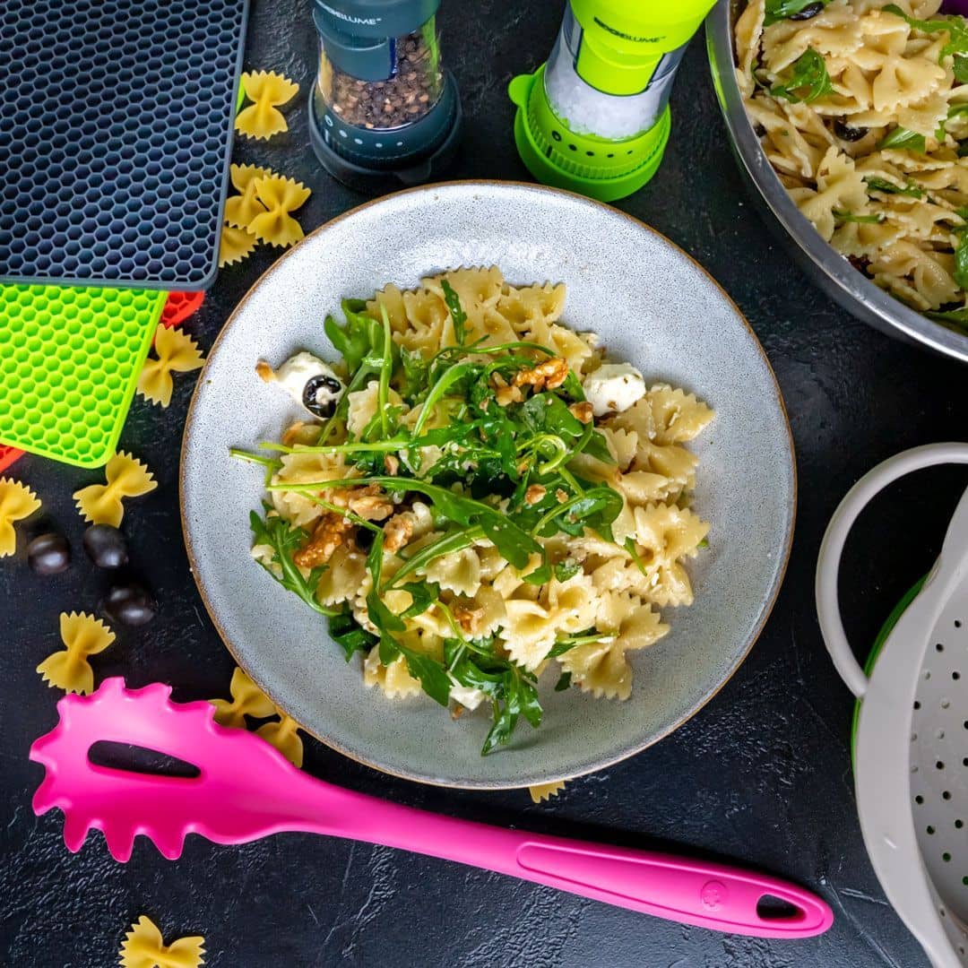 Eine Schüssel mit Farfalle-Nudeln, Rucola, Oliven, Käse und Walnüssen, umgeben von Küchenutensilien und Nudeln.