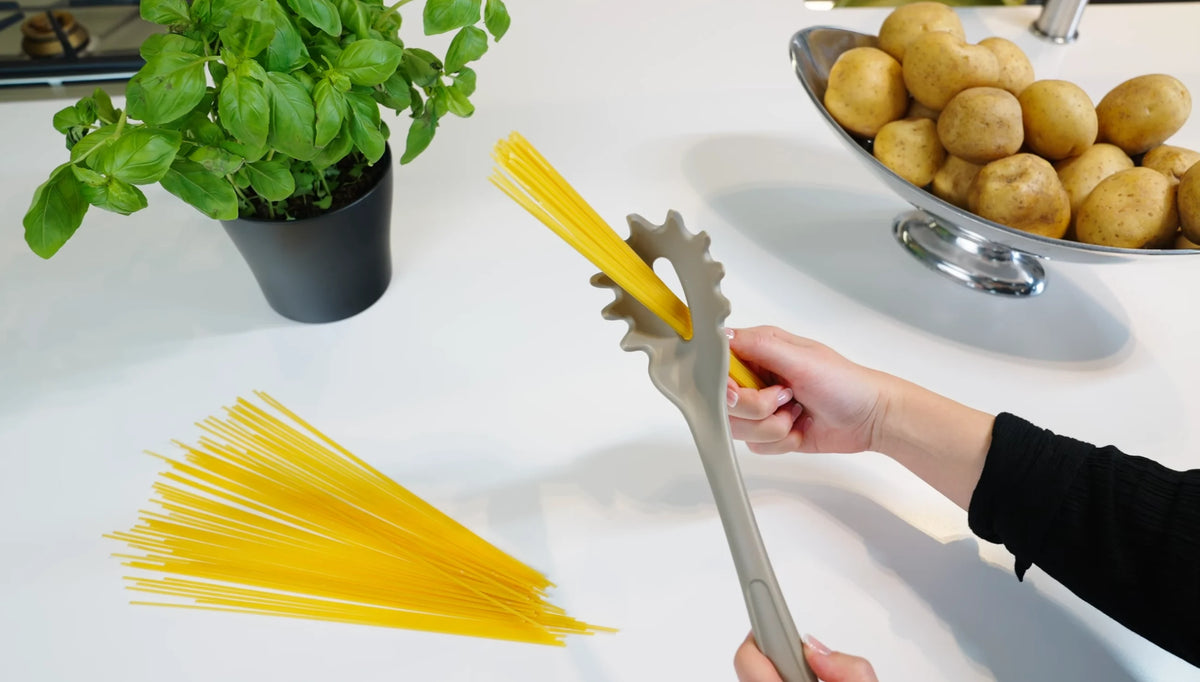 Eine Person misst mit einer Spaghettischaufel trockene Nudeln auf einem weißen Tisch ab, neben dem Basilikum und Kartoffeln liegen.