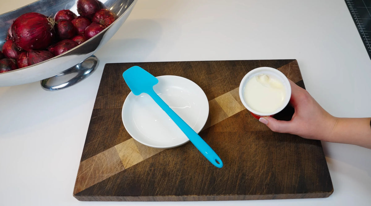 Eine Hand hält eine kleine Schüssel Joghurt neben einer leeren Schüssel, einem blauen Spatel und einem Schneidebrett auf einem Tisch.