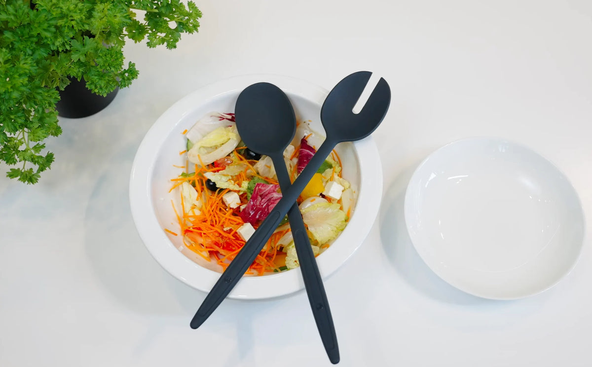 Weiße Schüssel mit Salat, darüber gekreuzte schwarze Servierbestecke, ein leerer Teller und daneben eine grüne Pflanze.