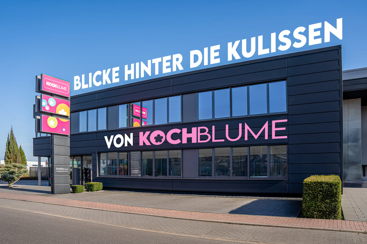 Modernes schwarzes Gebäude mit den Schildern „Blicke hinter die Kulissen“ und „Von Kochblume“, darüber ein klarer Himmel.