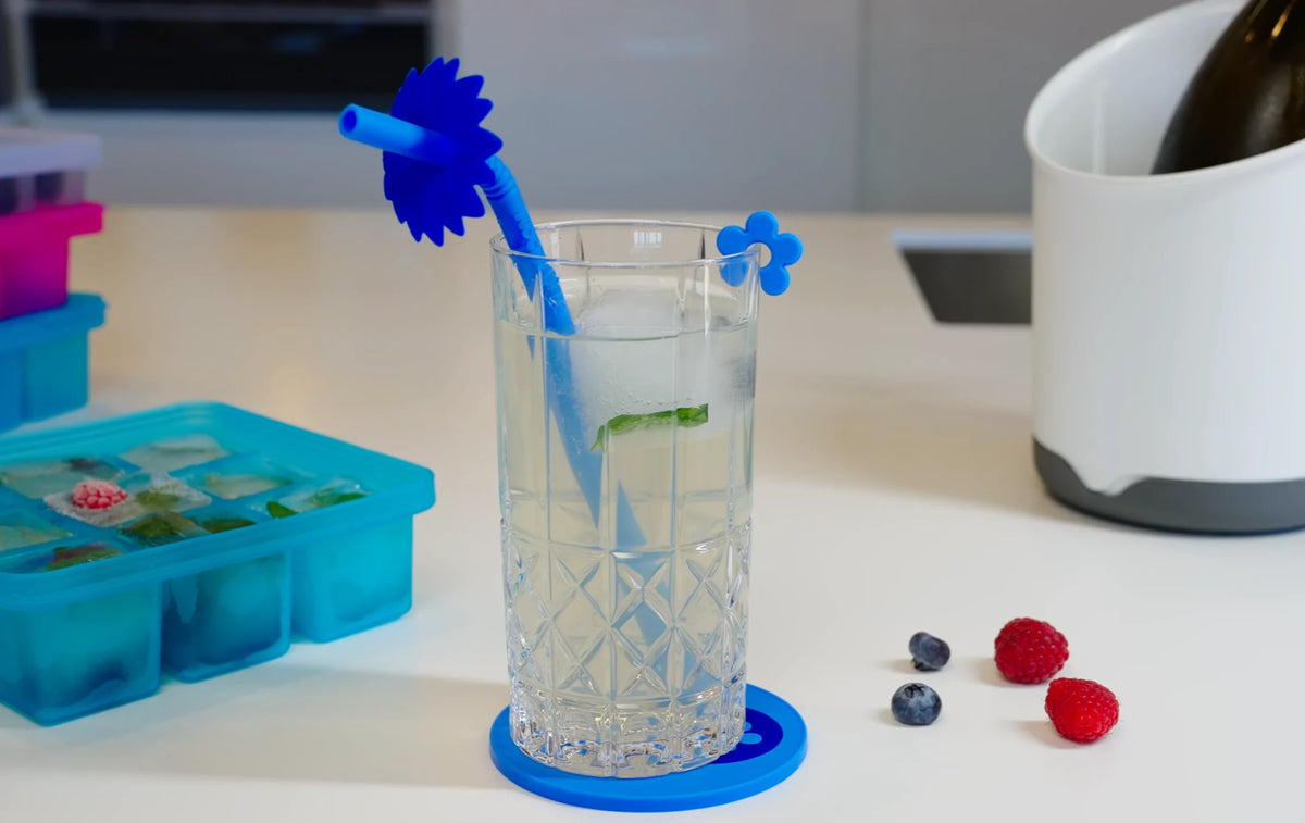 Ein Glas Eiswasser mit einem blauen Strohhalm, Beeren daneben und eine Eiswürfelform auf einer weißen Arbeitsplatte.
