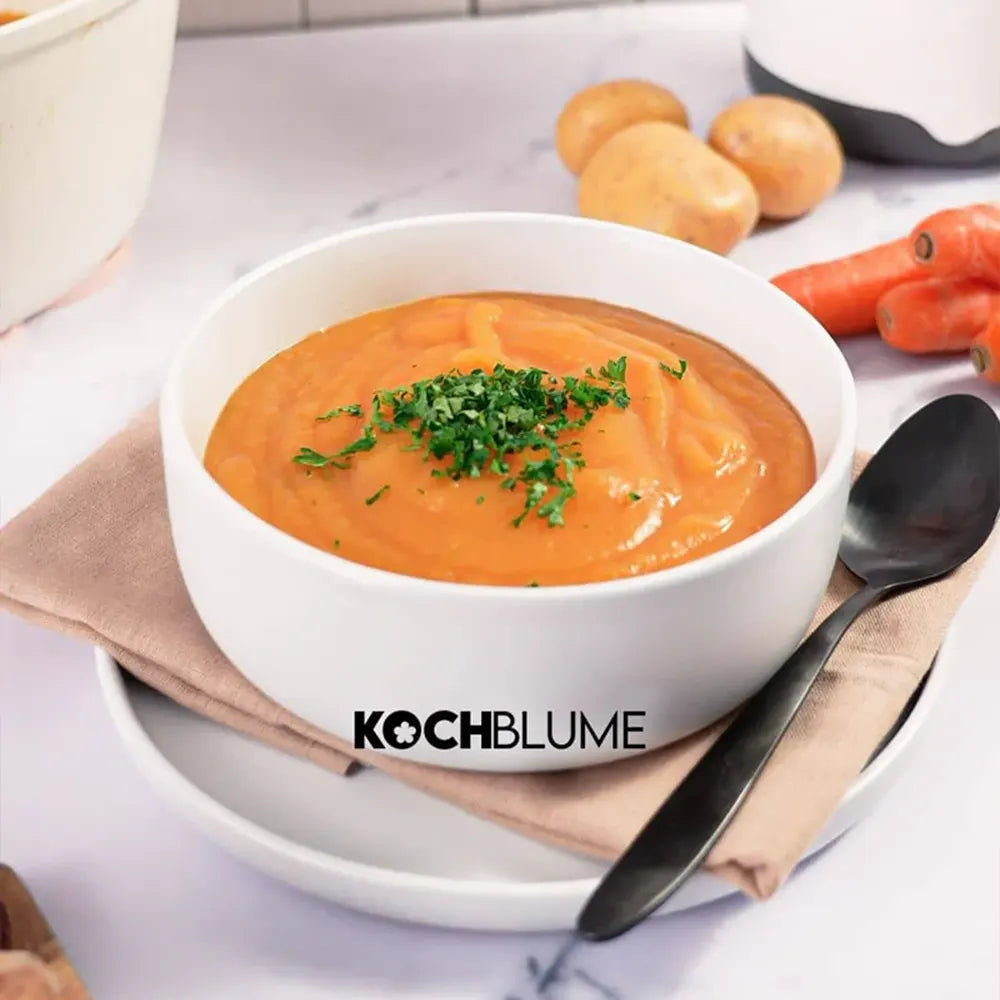 Eine Schüssel mit cremiger Orangensuppe, garniert mit Kräutern, auf einem Teller serviert, daneben ein Löffel sowie Kartoffeln und Möhren.