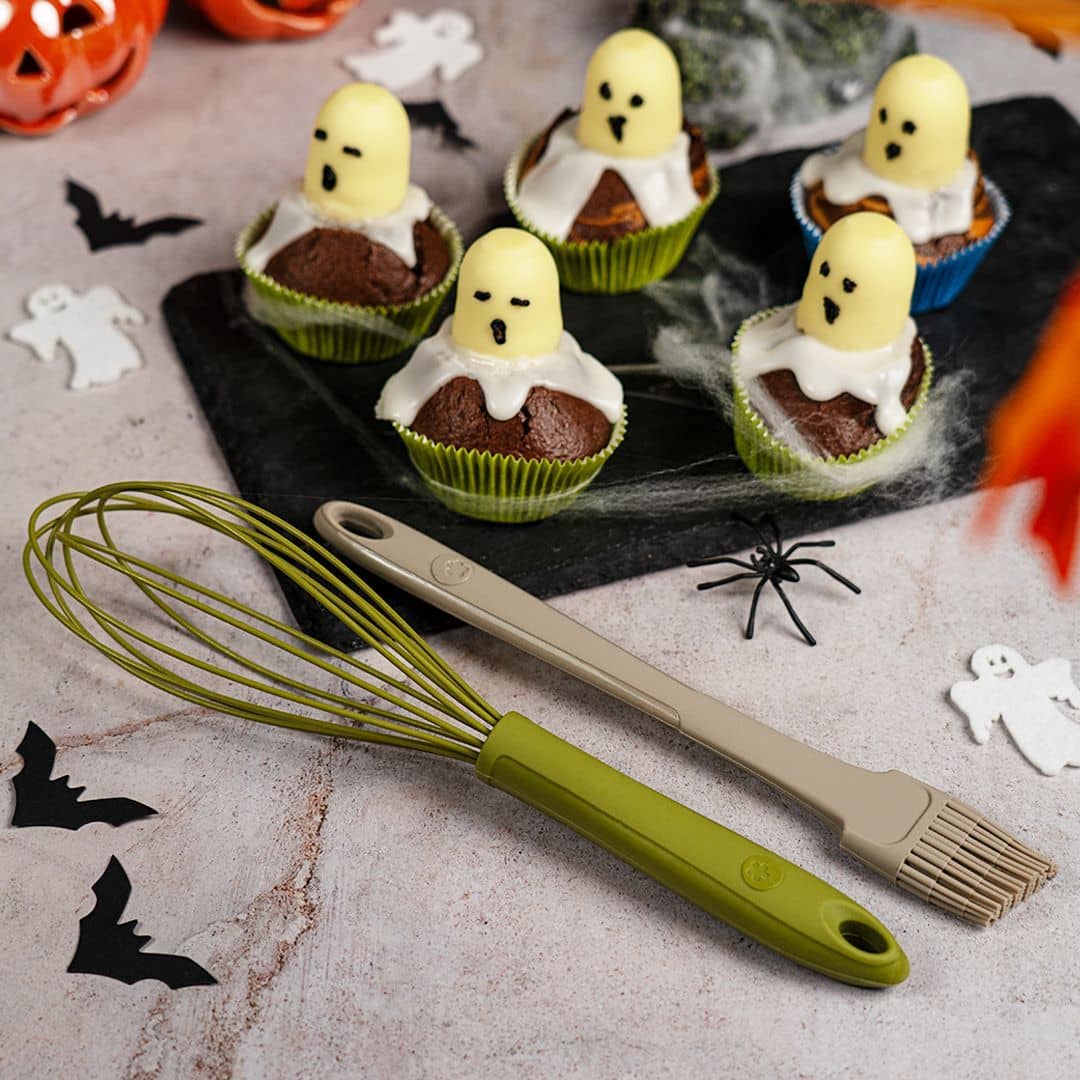 Fünf Geister-Cupcakes auf einer Schieferplatte, davor ein Schneebesen und ein Pinsel; drumherum Halloween-Dekoration.