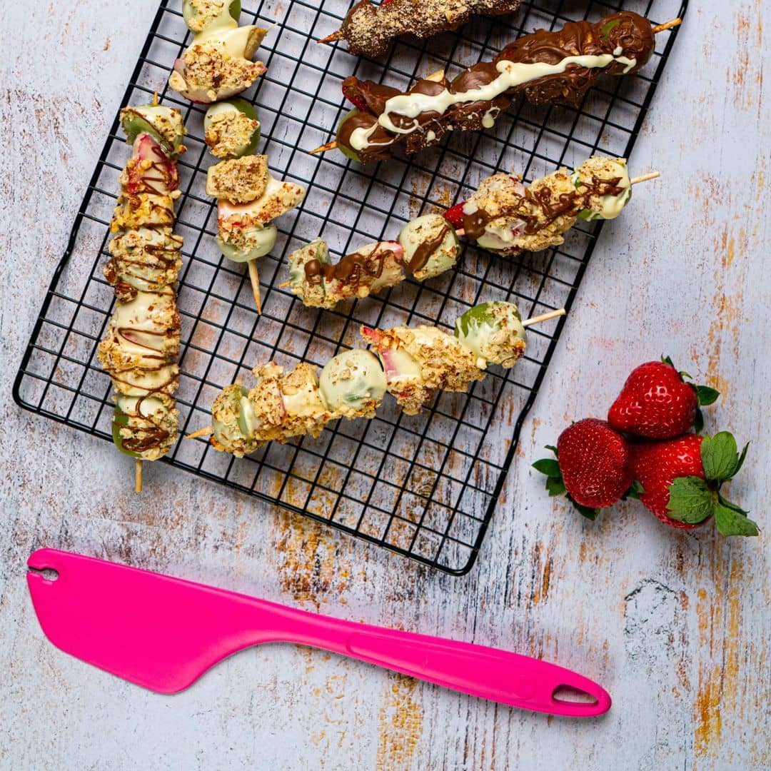 Mit Obst und Schokolade überzogene Spieße auf einem Abkühlgitter, frische Erdbeeren und ein rosa Spatel auf dem Tisch.