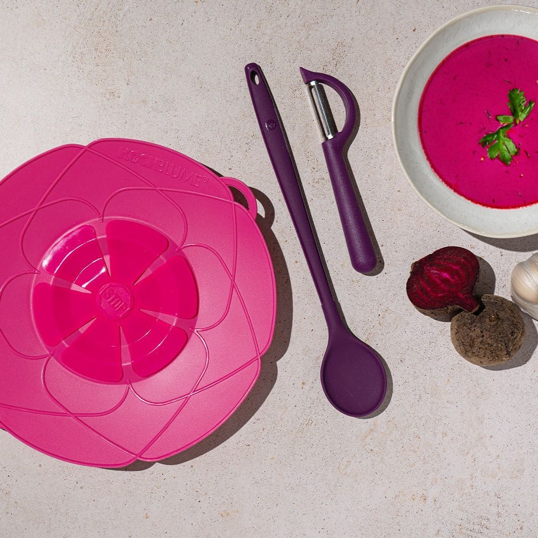 Rosa Silikondeckel, lila Löffel, Schäler, eine Schüssel mit Rote-Bete-Suppe und Gemüse auf einem hellen Untergrund.