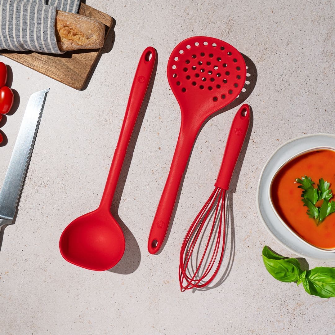 Drei rote Küchenutensilien neben einer Schüssel Tomatensuppe, Brot, einem Messer und frischen Basilikumblättern.