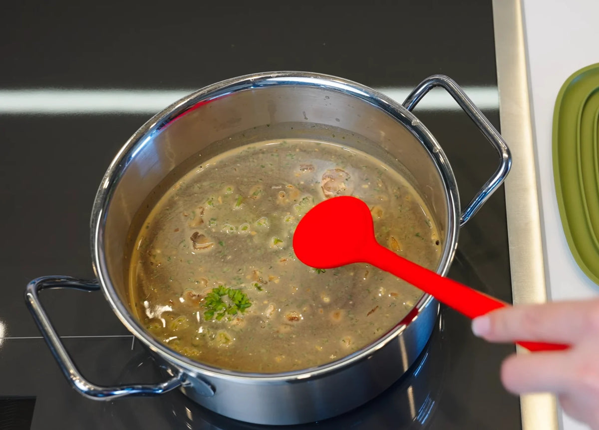 Eine Hand rührt mit einem roten Löffel in einem Edelstahltopf auf dem Herd in der Suppe um.